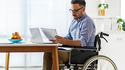 A man in a wheelchair, sitting at a desk in front of his laptop while holding some papers 