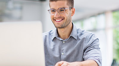 A man wearing glasses, smiling while using his laptop