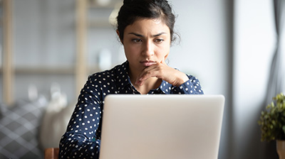 A woman in a dark blue polka dot blouse looking seriously at her open laptop.