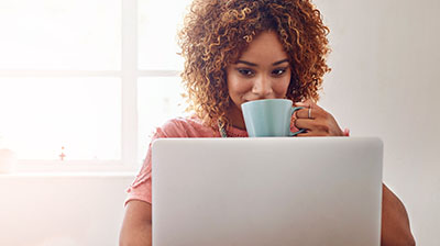 A woman drinking out of a light teal mug while looking at her laptop 