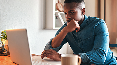 A man sitting at a desk and scrolling on his laptop while holding his chin 