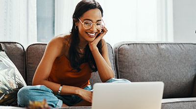 A woman sitting crisscross on a grey couch while smiling at her open laptop 