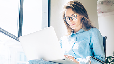 Woman in casual clothes sitting in an office chair, she has a laptop in her lap and she is looking down at the screen. 