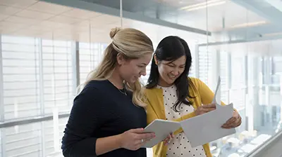 Two women standing next to each other while looking through stacks of papers in their hands 