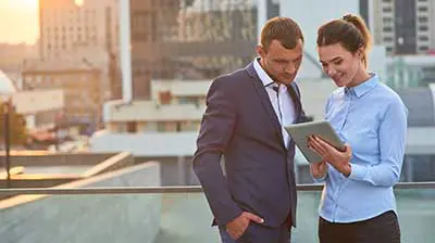 Man and a woman in professional attire on the roof of a building looking at a tablet