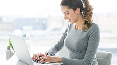 A woman in a grey sweater sitting at her desk smiling and typing on her laptop.