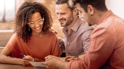 A woman and two men sit around a table holding pens and writing on the same piece of paper.