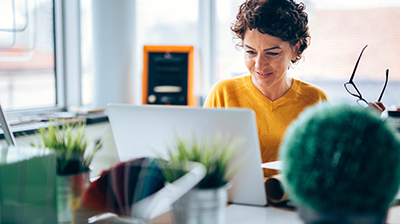 A woman holding her glasses in one hand and looking at her laptop while sitting at her desk