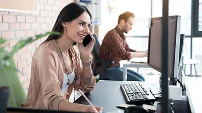 A woman working on a desktop computer while talking on a mobile phone