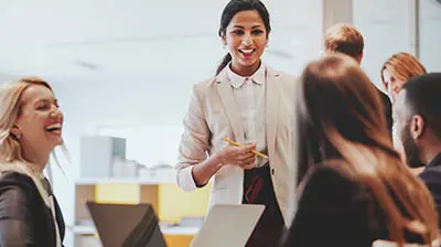A woman standing in front of three coworkers all sitting at a table while smiling at each other