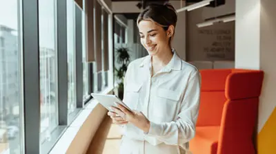A woman standing in front of a wall of windows, smiling down at the tablet in her hands. 