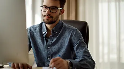 A man sitting at his desk looking at his desktop monitor while holding a pen over a notepad 