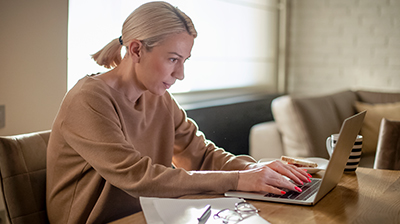 A woman sitting at her dining table typing on a laptop while a plate of toast and a mug sits on the table beside her 
