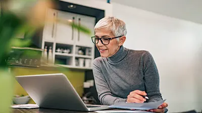 A woman in a gray turtleneck holding a paper and pointing a pen over the paper while leaning over her laptop 