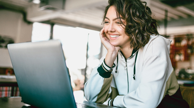 A woman in a white hoodie smiling at her laptop with a hand on her cheek