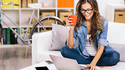 Woman sitting on couch holding coffee mug and using a laptop.
