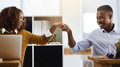 A man and a woman fist bumping each other while each sitting at their own desks 