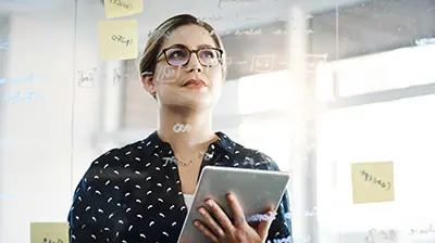 A woman holding a tablet while looking up at a transparent whiteboard with writing on it 