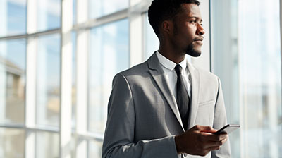 Man in business attire looking out a window while holding a cell phone in his hand.