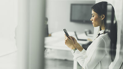 Woman in casual attire standing in an office while holding a mobile phone in front of her.