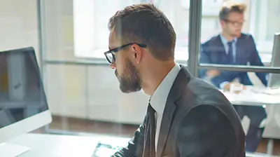 A man in glasses looking at his computer, coworker in the background