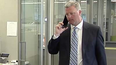 Man dressed in business attire walking through an office while holding a phone to his ear.