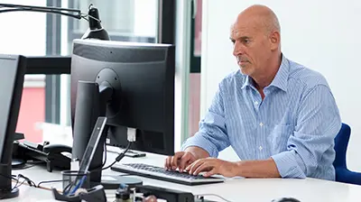 Man in casual attire sitting at a desk in an office with two large computer screens in front of him. 