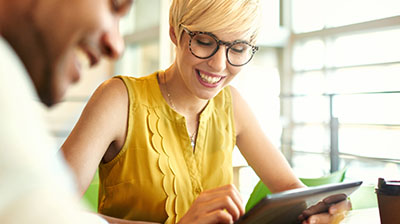 Smiling woman in casual attire with glasses looking down at a tablet in her hands, there is a man sitting in front of her to the left of the image, also smiling.