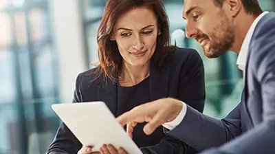 A man and woman in business attire sitting at a table, they are both looking down at a tablet device in the woman's hands.