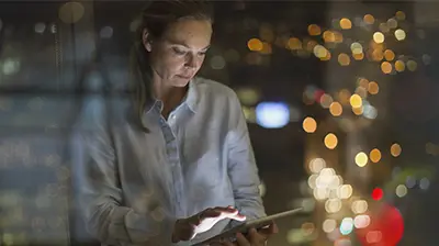 Woman in casual attire standing in front of a window looking down at a tablet in her hands.