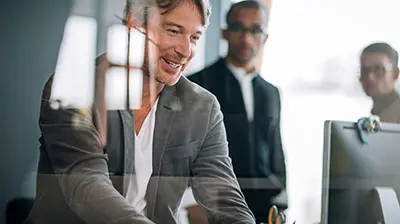 Three men in business attire in an office, one man is in front looking down at a computer screen.