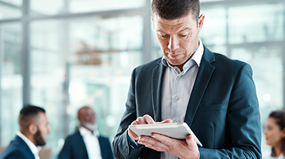 Man in business attire standing in front of colleagues conversing while he uses a tablet.