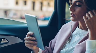 Woman in business attire sitting in car with window down using a tablet with earphones in her ears.