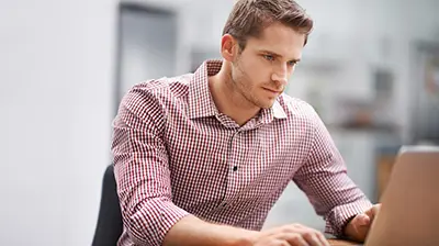 Man in casual attire sitting at a desk typing on an open laptop in front of him. 