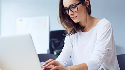 Woman in business attire sitting at a desk, she is looking down at an open laptop in front of her.