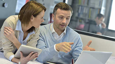Man and woman sitting at desk with laptop; Man gestures towards laptop screen while woman looks on and holds a tablet