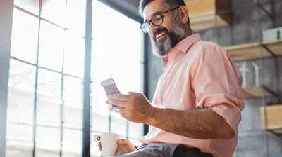 Business man in a modern office looking at his phone while drinking coffee.