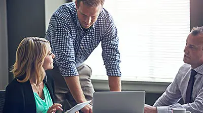 A man and woman are sitting and talking with a laptop between them. Another man is standing between them looking at the laptop. The woman is holding a tablet