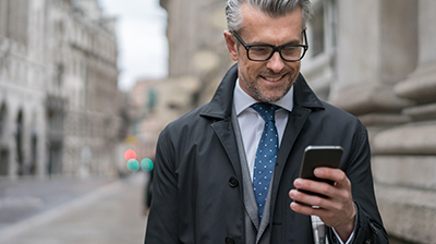 A businessman wearing glasses, standing outside, looking at his mobile phone